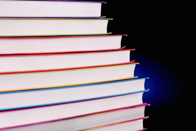 A stack of thin books of same sizes, with white pages and colorful flexicovers arranged on a semi rainbow order. They are standing in the dark, in front of a black background lit by a small deep blue spot.